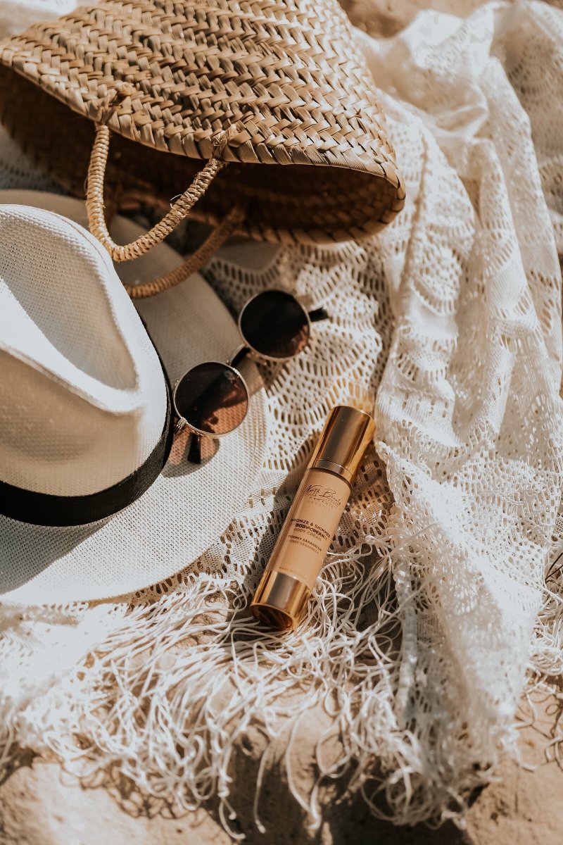 Tinted Glow Cream i gyllene tube med pump ligger bland vita textilier, solglasögon och korgväska i naturlig belysning