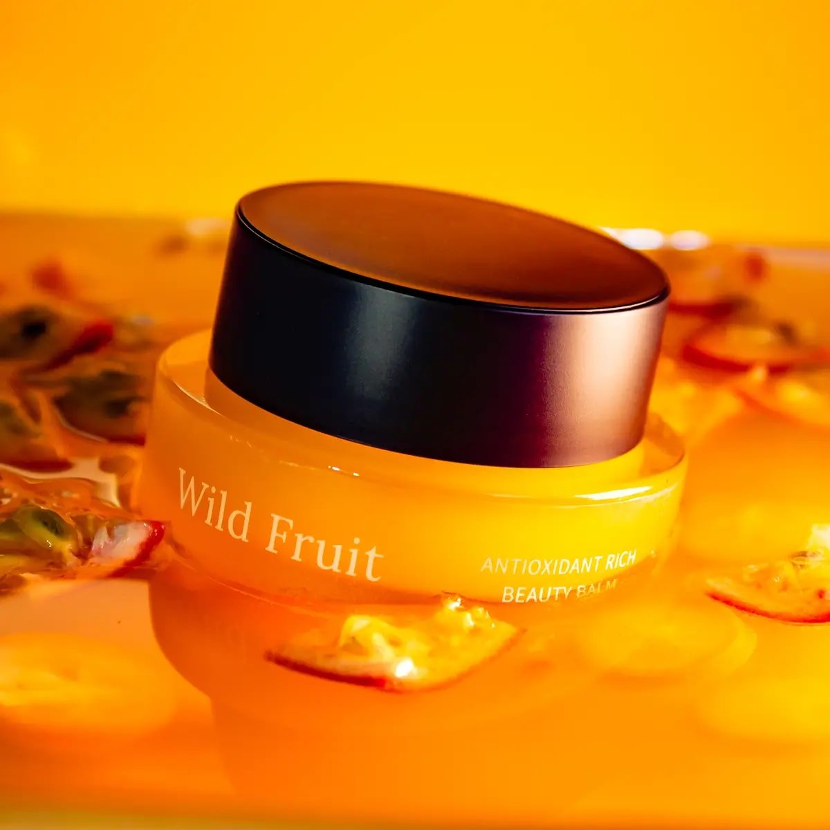 Usva Wild Fruit Beauty Balm i orange glasburk med svart lock, omgiven av torkade fruktskivor mot gul bakgrund