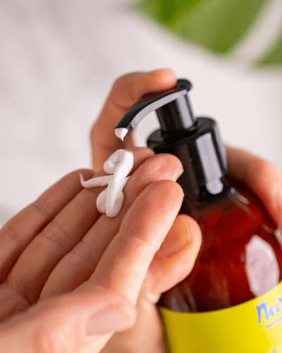 Händer som pumpar ut vit lotion från brun glasflaska med gul etikett - Wild Lemongrass Hand och Body Lotion 300ml