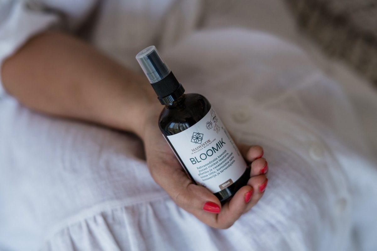 Mörk glasflaska med spraytopp innehållande Kropps- och Massageolja 2i1, 100ml hålls i hand med röda naglar