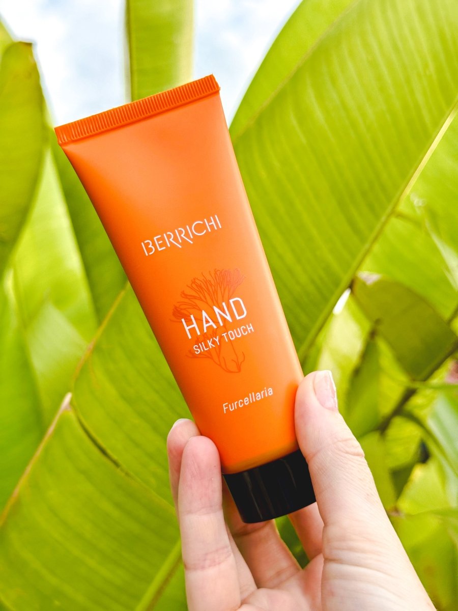 Orange handkrämstub från Berrichi med vit text, hålls i hand mot gröna tropiska blad i bakgrunden