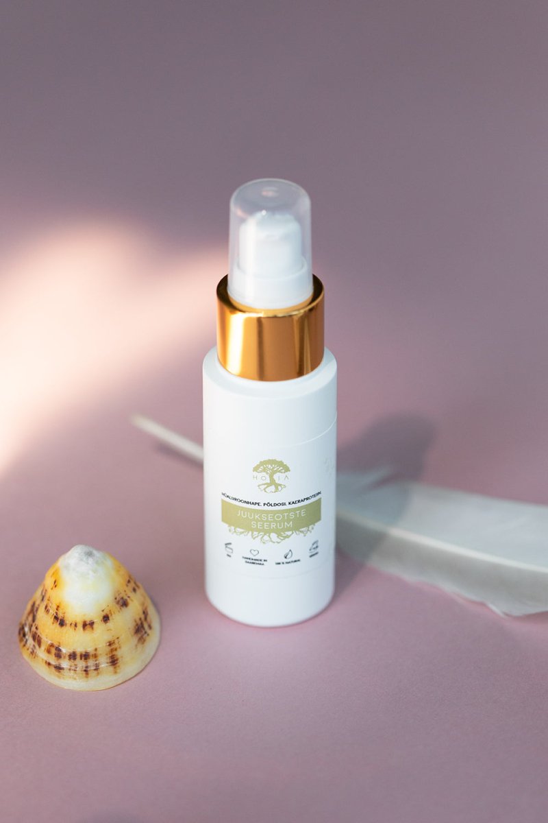 HOIA Hair Ends Serum 50ml i vit flaska med gulddetaljer och pump, placerad bredvid snäckskal på rosa bakgrund
