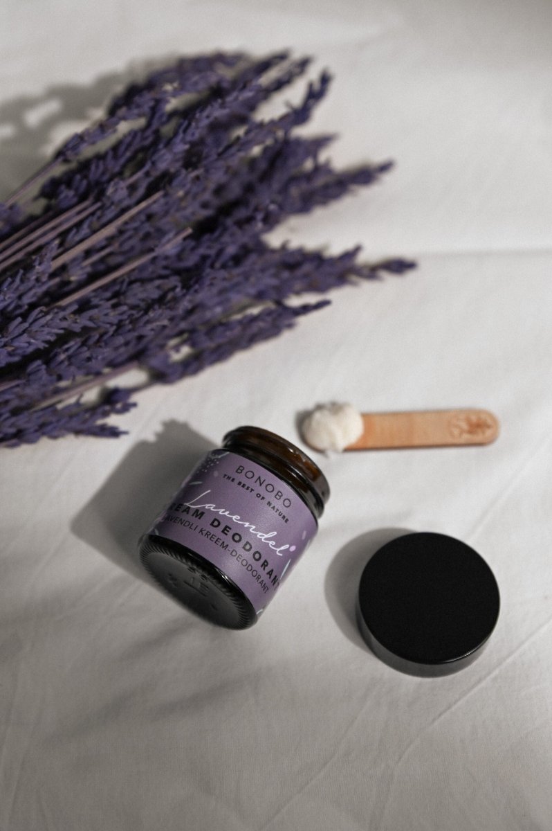 Deodorantkräm Lavendel i glasburk med lila etikett och svart lock, bredvid torkad lavendel och träsked på vit bakgrund