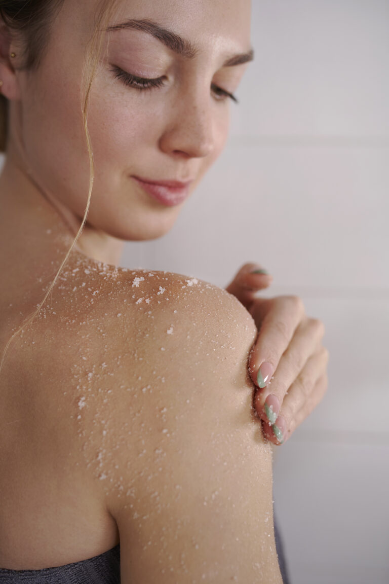 Kvinna applicerar Lavendel Kroppspeeling med Grapefrukt på axel - vita exfolierande korn syns på huden