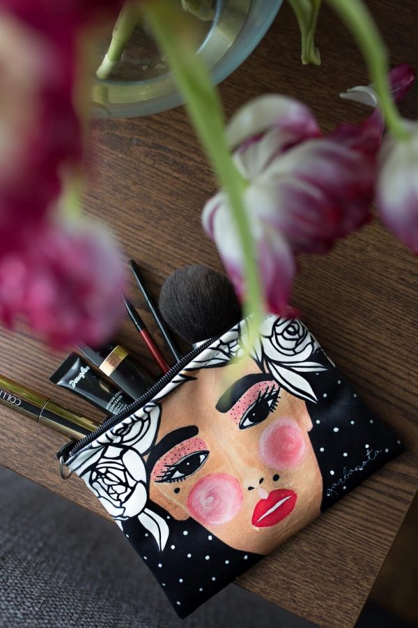 MAKEUP BAG YOKO BLACK med illustrerat kvinnoansikte och vita rosor, fylld med sminkborstar på träbord