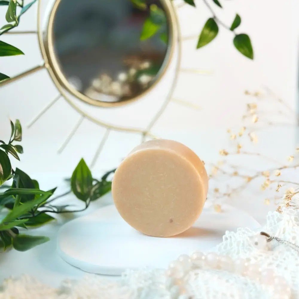 Rund beige ansiktsrengöringsbar med rosa lera framför spegel och gröna växter på vit yta