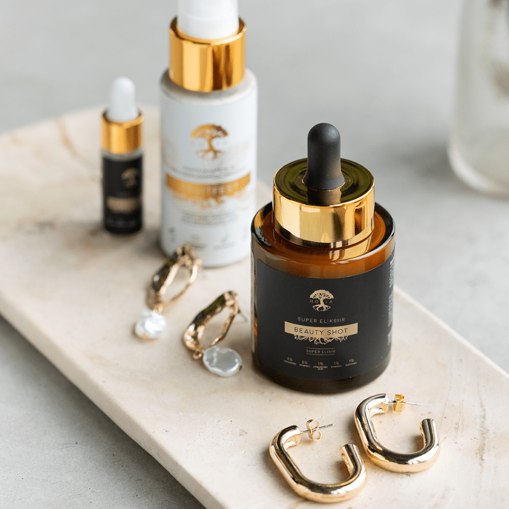 Beauty SHOT Serum 30ml i mörk glasflaska med gulddetaljer och pipett, omgiven av hudvårdsprodukter och smycken