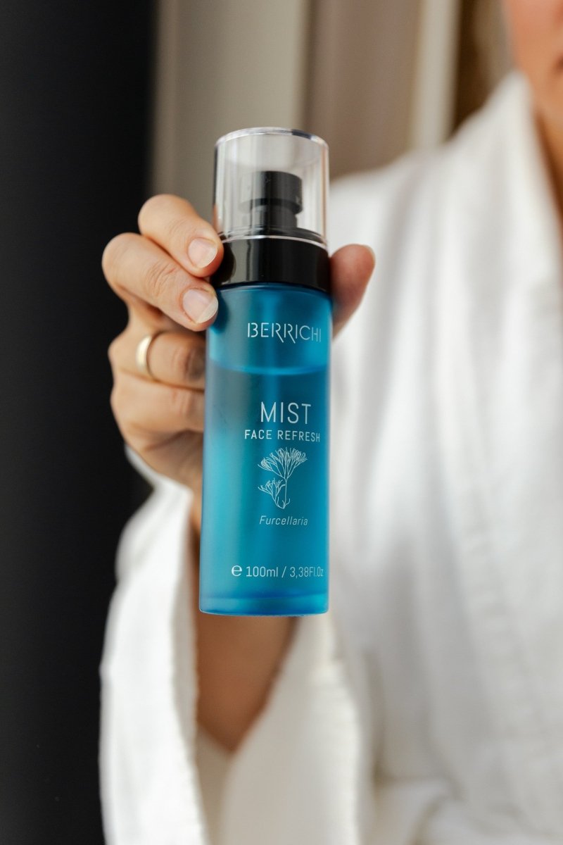 Blå glasflaska med spraypump innehåller BERRICHI MIST Face Refresh 100ml, hålls av hand i vit skjorta