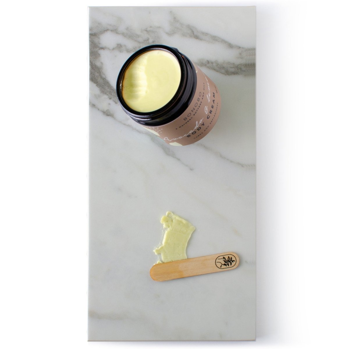 Öppen rosa burk med gul Body Cream - Avokado och Citron på marmorbakgrund, med träspatel och produktprov