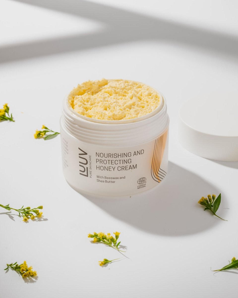 Öppen vit burkförpackning med gul närande honung body cream, omgiven av små gula blommor på vit bakgrund