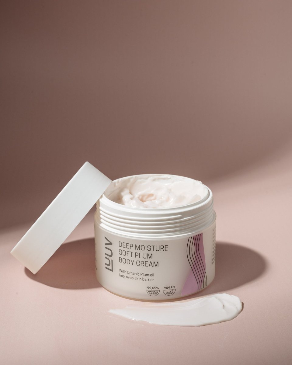 Öppen vit burk med Body Cream Plommonolja, krämig vit textur synlig, lock bredvid, rosa bakgrund