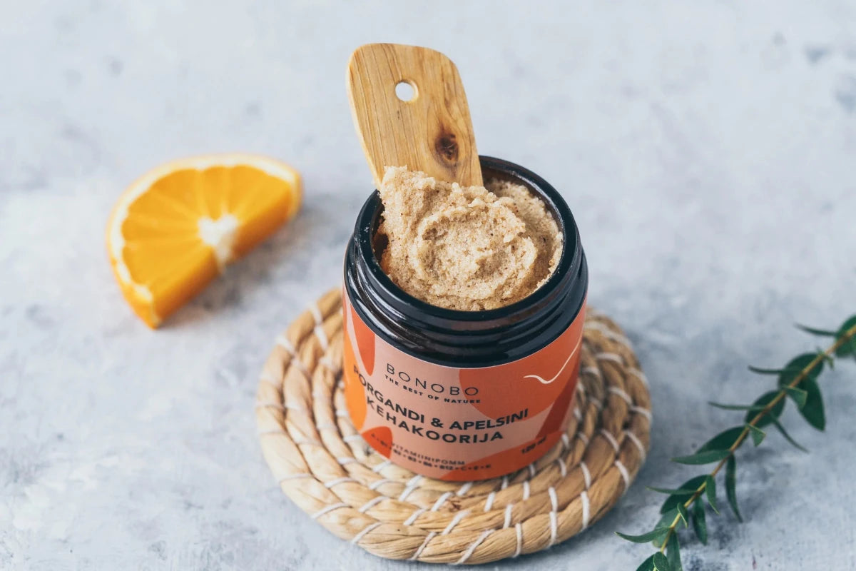 Bonobo Body Scrub med apelsin och morot i orange burk med svart lock, träsked med beige scrub-textur, apelsinskiva bredvid