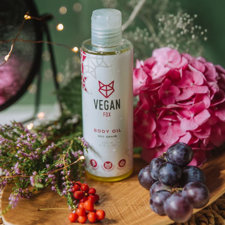 Red Grape Body Oil i genomskinlig flaska med vit etikett och rött foxlogotyp, omgiven av rosa blommor och druvor