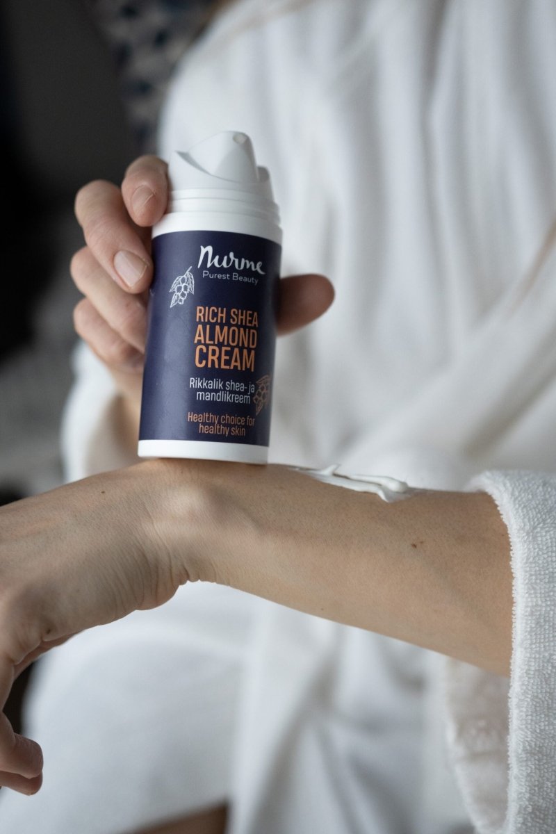 Ekologisk Shea & Mandelkräm i vit pump-flaska med mörkblå etikett hålls av hand, vit kräm synlig på underarm