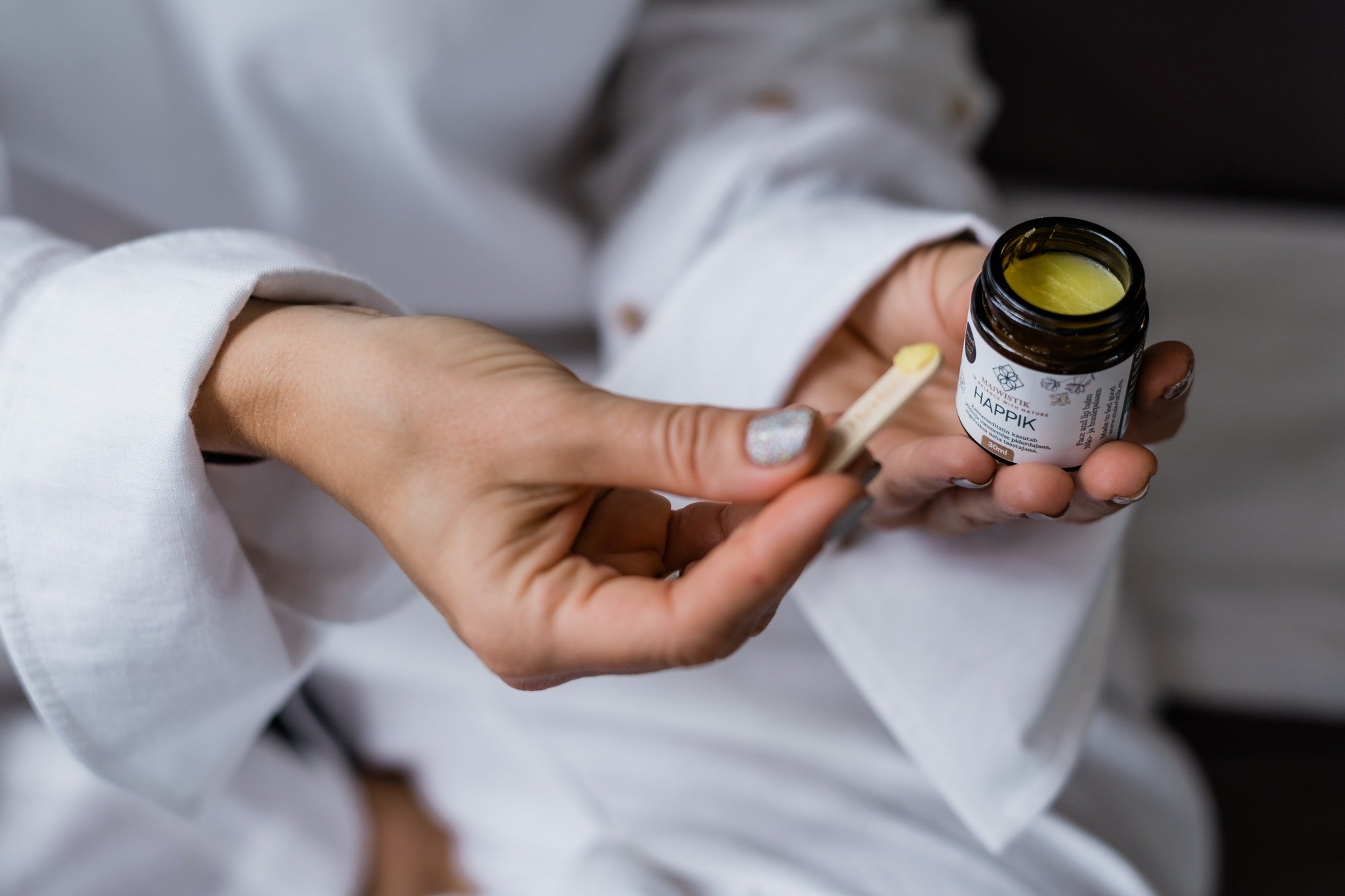 HAPPIK Rejuvenating Face Balm i mörk glasburk med gul balsam, hålls i hand med träsked för applicering