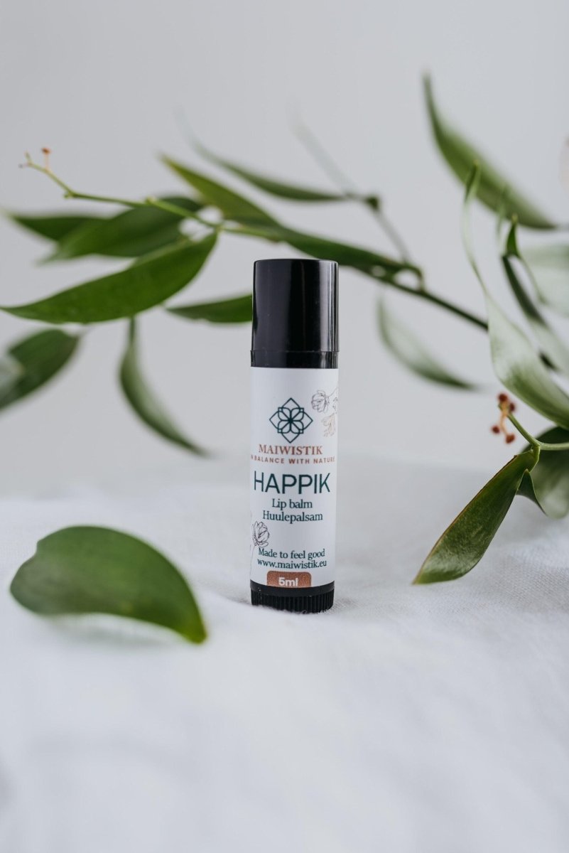 Happik naturlig läppbalsam i vit cylinderförpackning med svart lock, 5ml, omgiven av gröna växtblad på vit bakgrund