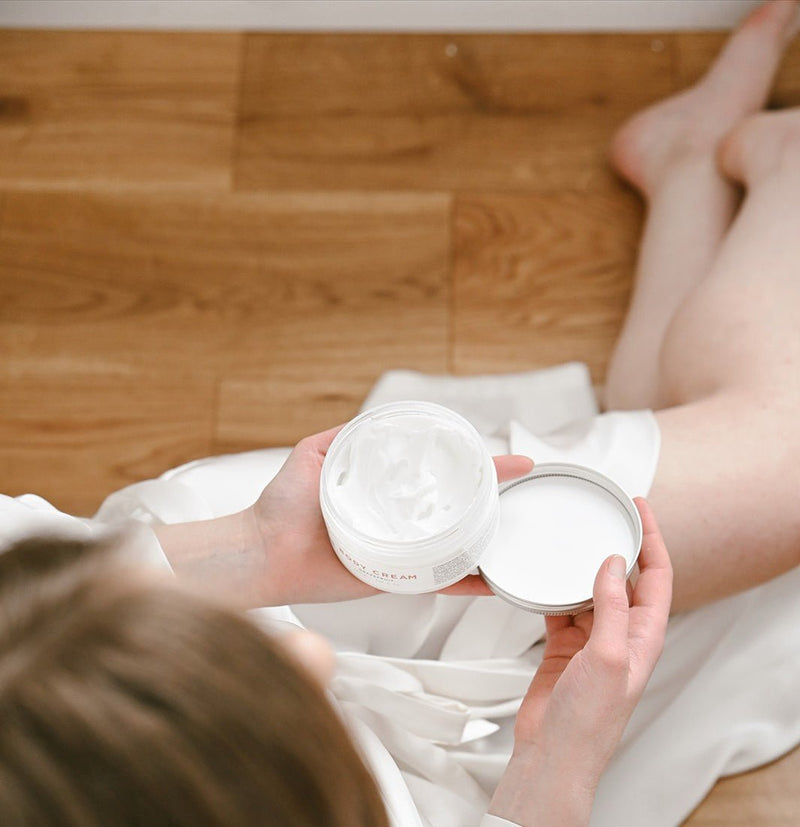 Händer håller öppnad burk med Lindblomma Body Cream – vit kroppskräm för torr hud mot träbjälkegolv