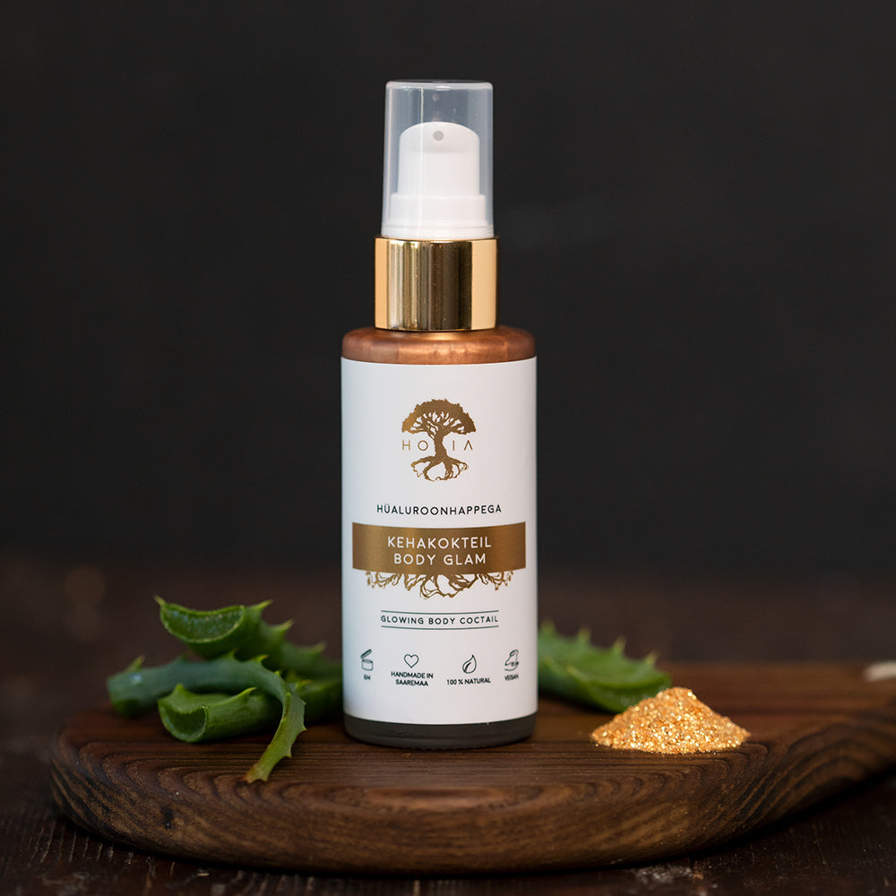 Kroppslotion Body Glam i vit sprayflaska med gulddetaljer på träunderlägg med aloe vera och guldiga partiklar
