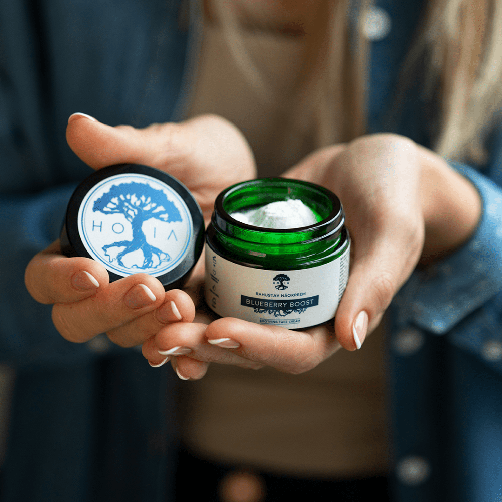 Lugnande Ansiktskräm Blåbär Boost 30ml i grön glasburk med vit kräm, hålls i händer med öppet lock som visar blått trädlogo