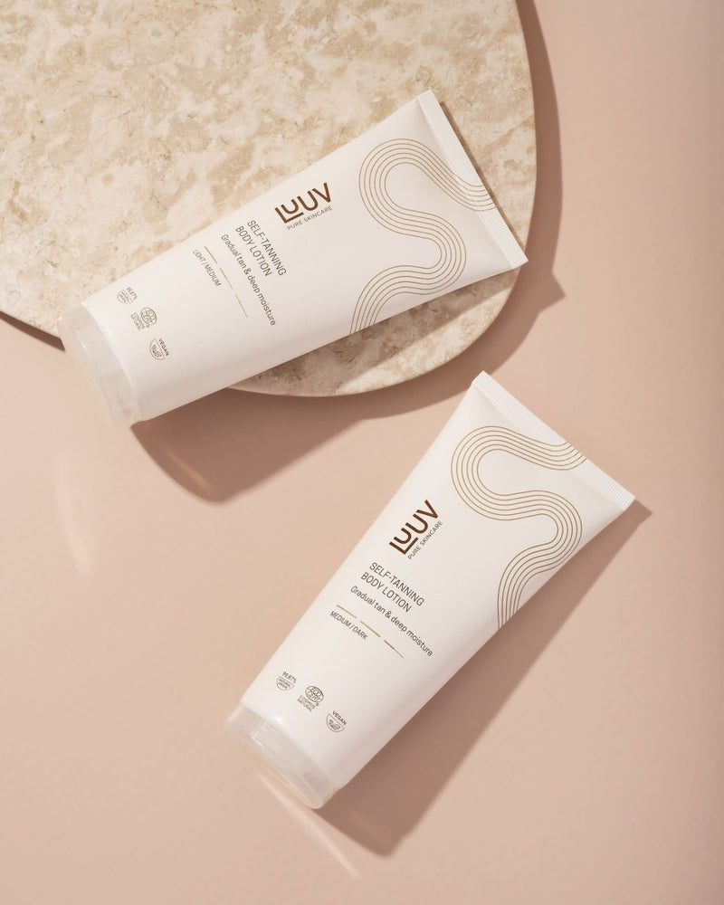 Två vita tuber med LUUV Brun Utan Sol Body Lotion, elegant design med bruna vågor, placerade på beige yta vid marmorbricka