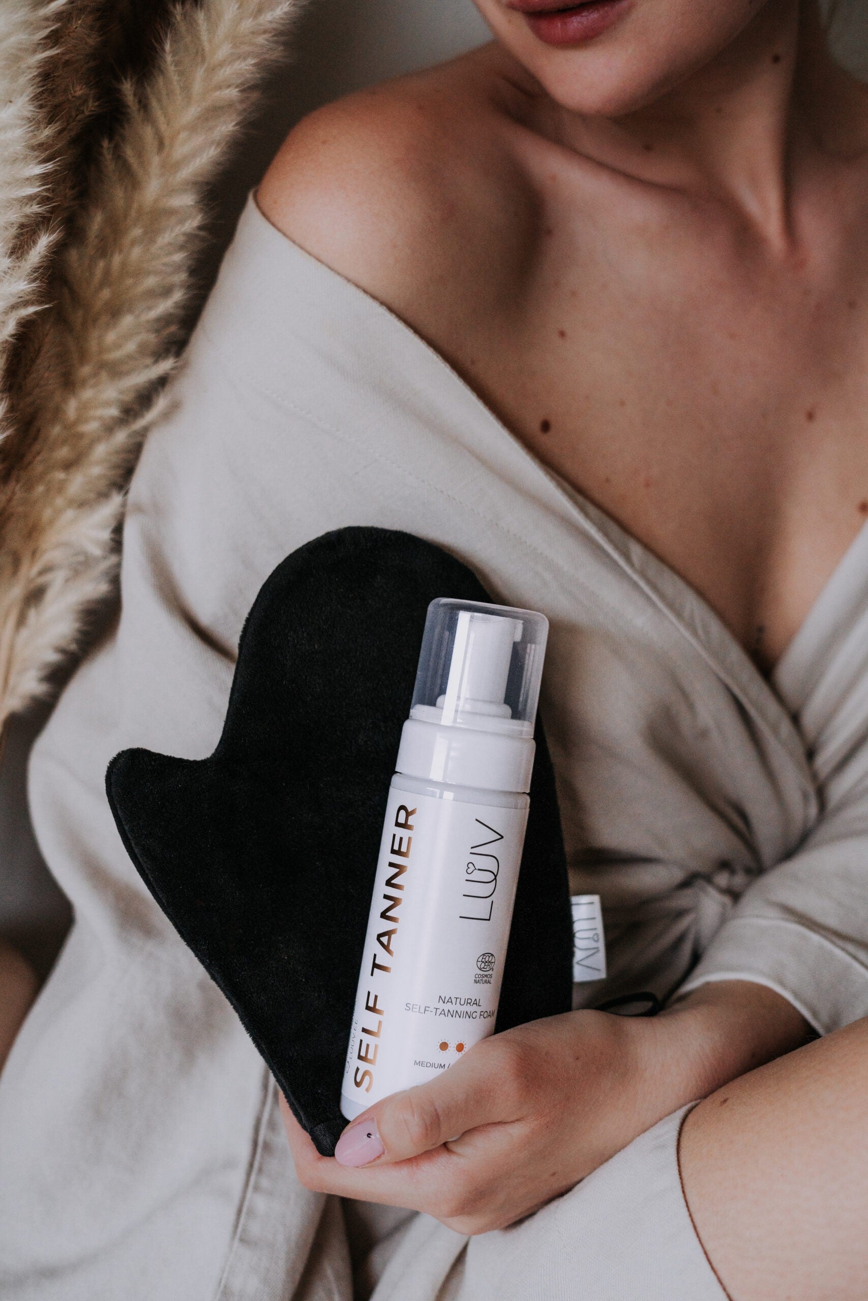 Kvinna i beige morgonrock håller en vit flaska LUUV Natural Self-Tanning Foam tillsammans med en svart appliceringshandske