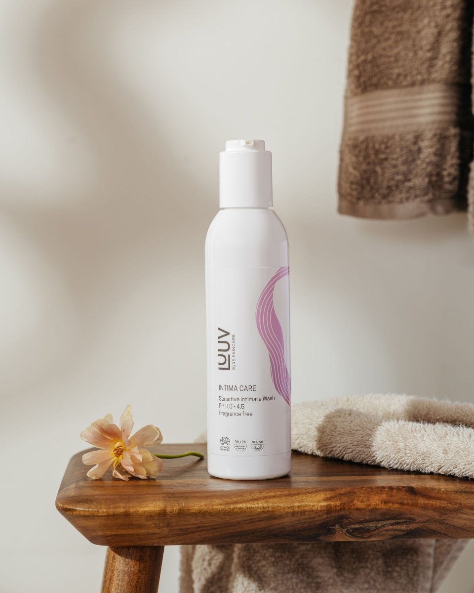 Vit flaska med pump innehållande LUUV Sensitive Intimate Wash 200ml, rosa design, placerad på träbänk med blomma