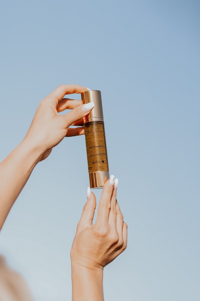 Natural Tanning Oil 50ml i elegant gyllene flaska med pump, hålls av händer mot ljusblå himmel