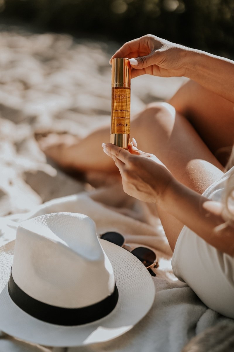Natural Tanning Oil 50ml i gyllene flaska med guldlock hålls i händerna, solljus på hud och vit hatt