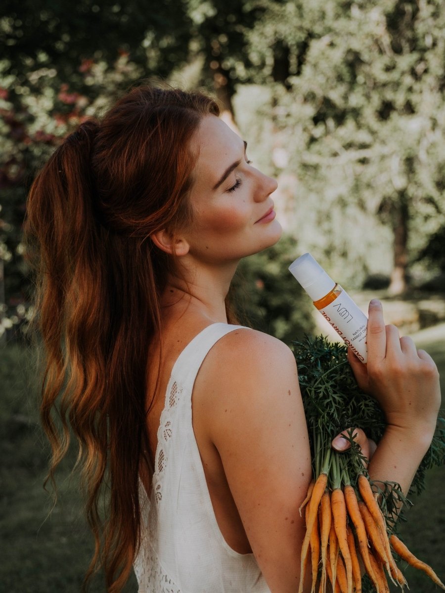 Kvinna håller vit flaska med Naturlig Carrot Dry Oil med Aloe Vera 100ml och färska morötter i trädgård