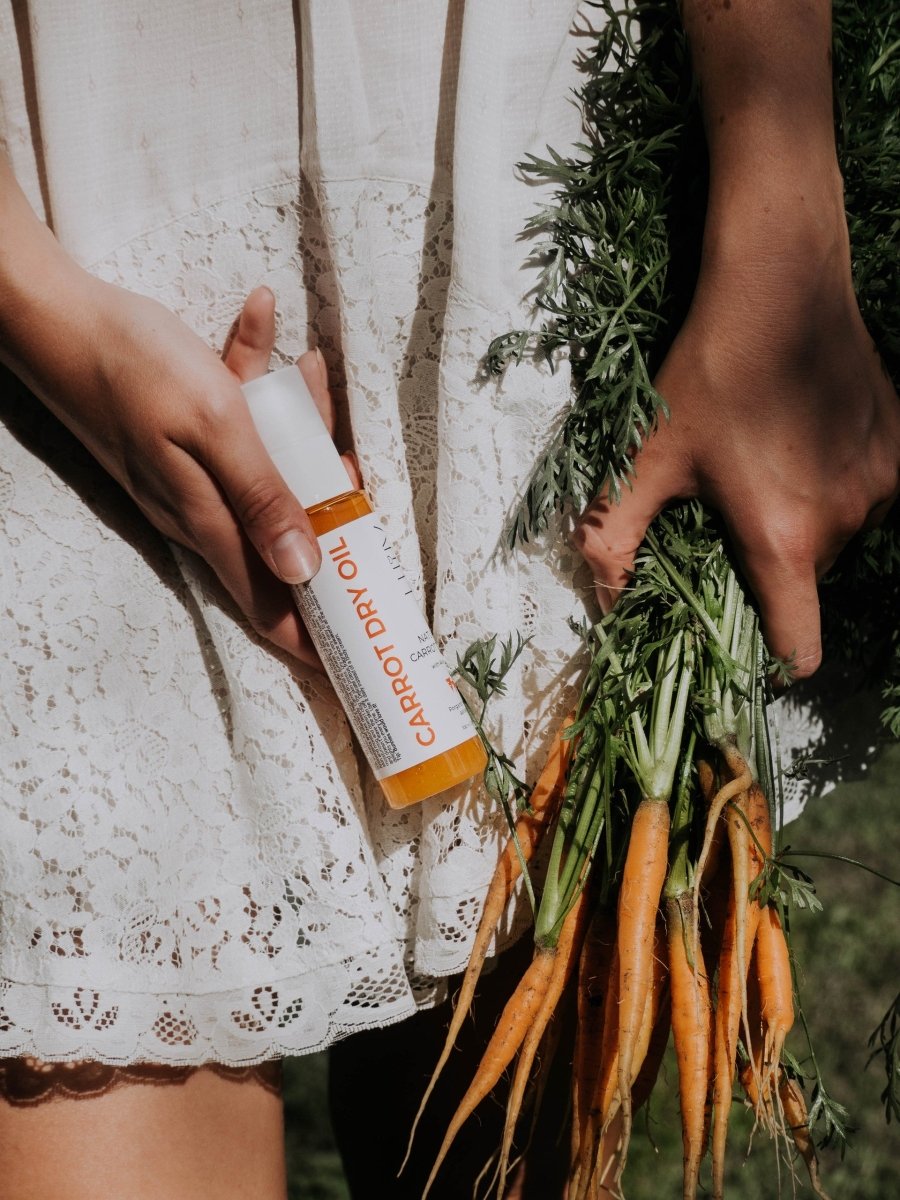 Vit flaska med orange detaljer Naturlig Carrot Dry Oil med Aloe Vera 100ml hålls av hand bredvid färska morötter