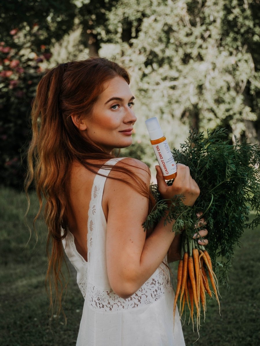 Kvinna håller vit sprayflaska med orange detaljer - Naturlig Carrot Dry Oil med Aloe Vera bland gröna morötter