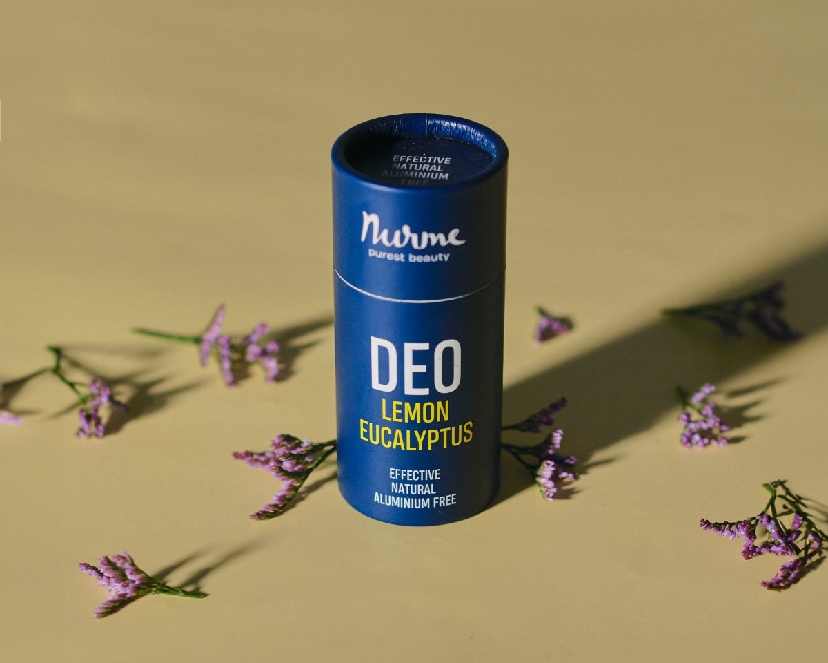 Blå cylinderformad deodorant från Norme med texten DEO LEMON EUCALYPTUS, omgiven av lila blommor på beige bakgrund