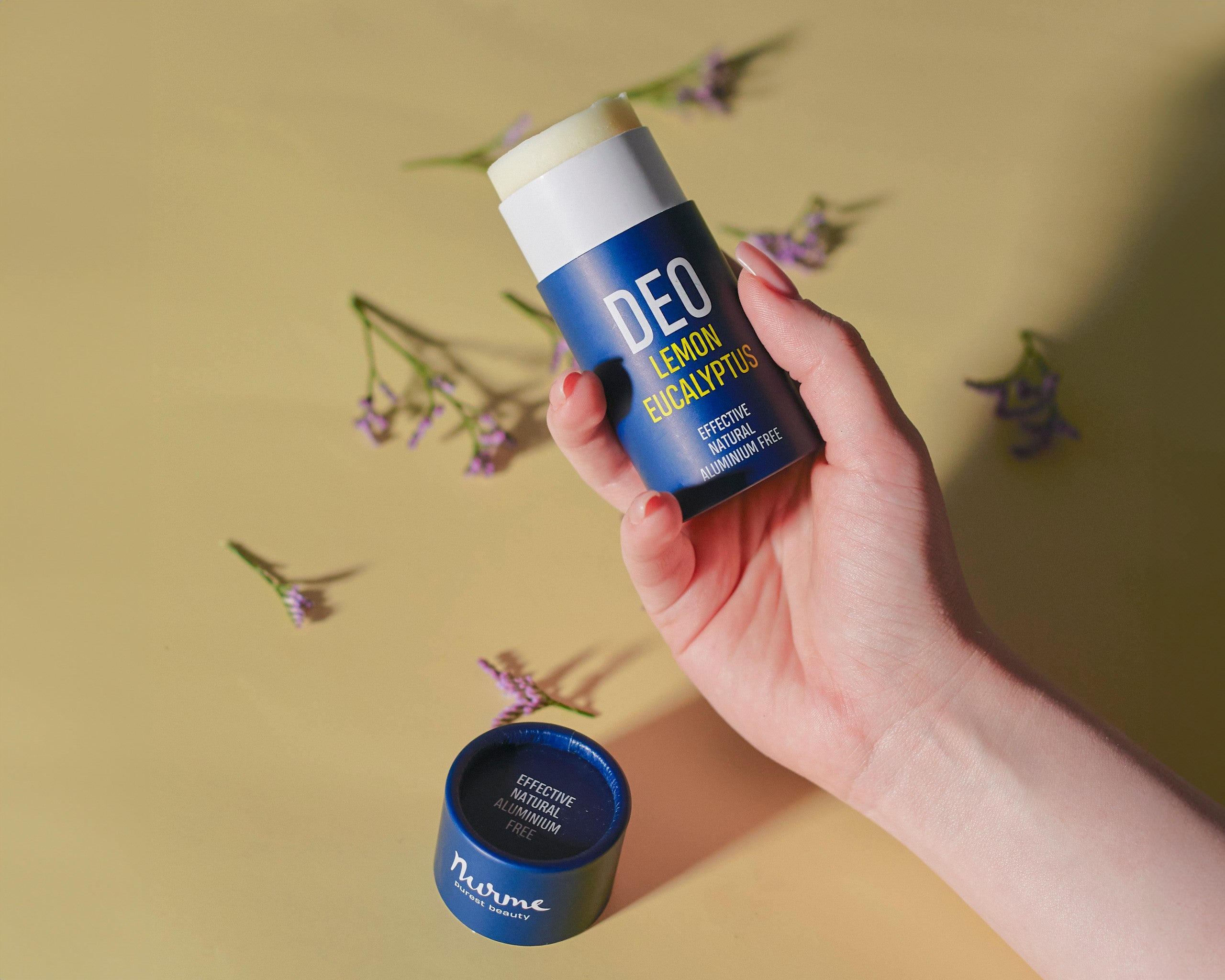 Hand håller upp Nurme naturlig deodorant citron och eukalyptus med öppnat lock framför, omgiven av lila blommor