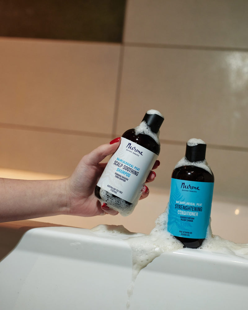 Nurme Balneological Peat Strengthening Conditioner i blå etikett på svart flaska, omgiven av bubblor i badkar