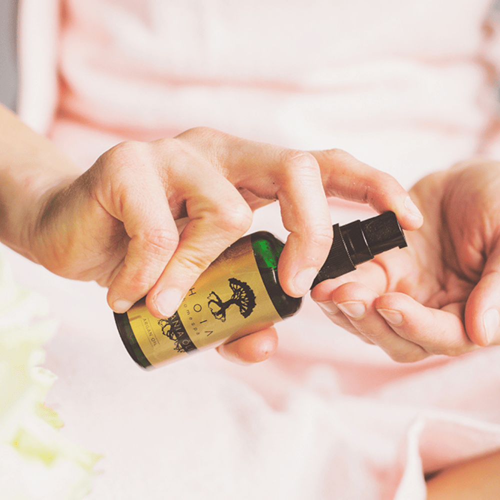 Kvinna applicerar Organic Argan Oil från gul flaska med svart pump i handflatan mot ljus bakgrund