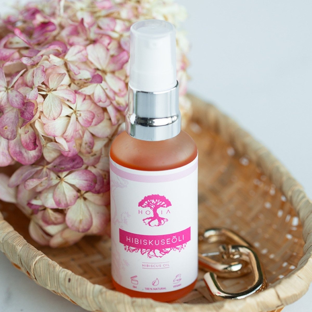 Organic Hibiscus Beauty Oil 50ml i glasflaska med vit etikett och rosa logotype, silvrig pump, bredvid torkade blommor