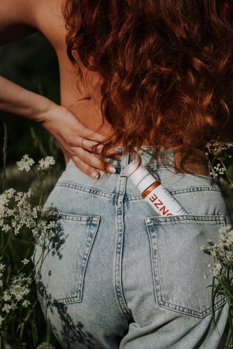 Kvinna med lockigt hår tar upp Shimmering Carrot Dry Oil Bronze från bakfickan på ljusblå jeans, omgiven av vita blommor