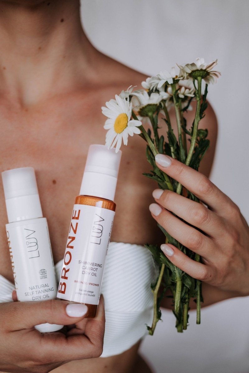 Shimmering Carrot Dry Oil Bronze i vit flaska med orange detaljer hålls bredvid vita prästkragar, naturlig hudvård