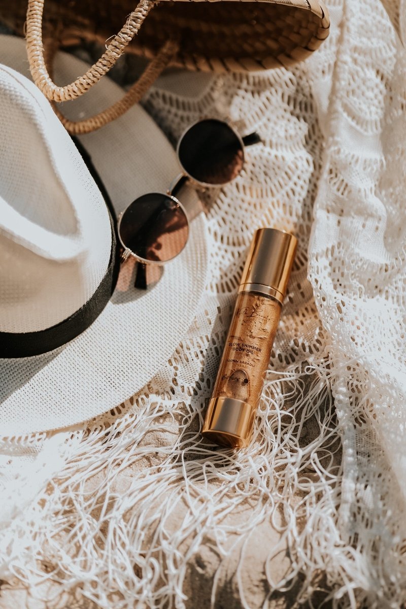 Glow Oil 50ml i gyllene flaska med pump, omgiven av solglasögon, korg och vita textilier i naturlig belysning